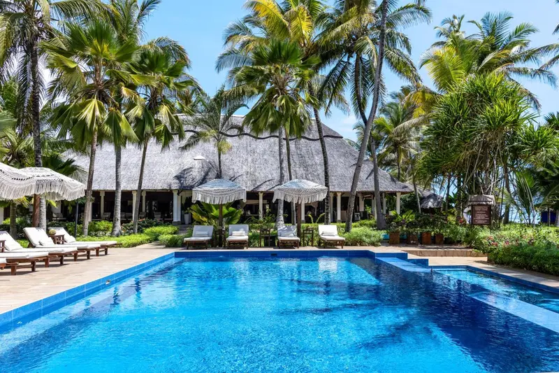 Une piscine turquoise bordée de transats, devant un bâtiment à toit de chaume et une végétation luxuriante.