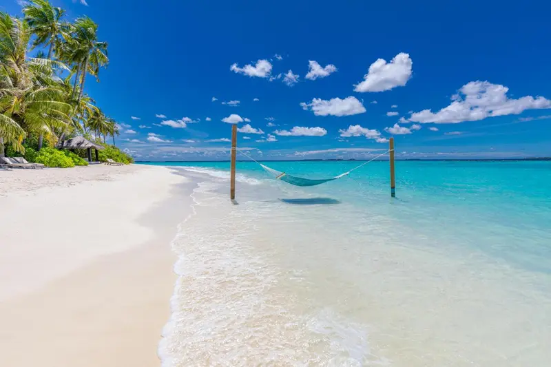 Un hamac est tendu entre deux poteaux sur une plage de sable blanc bordée d'eau turquoise.