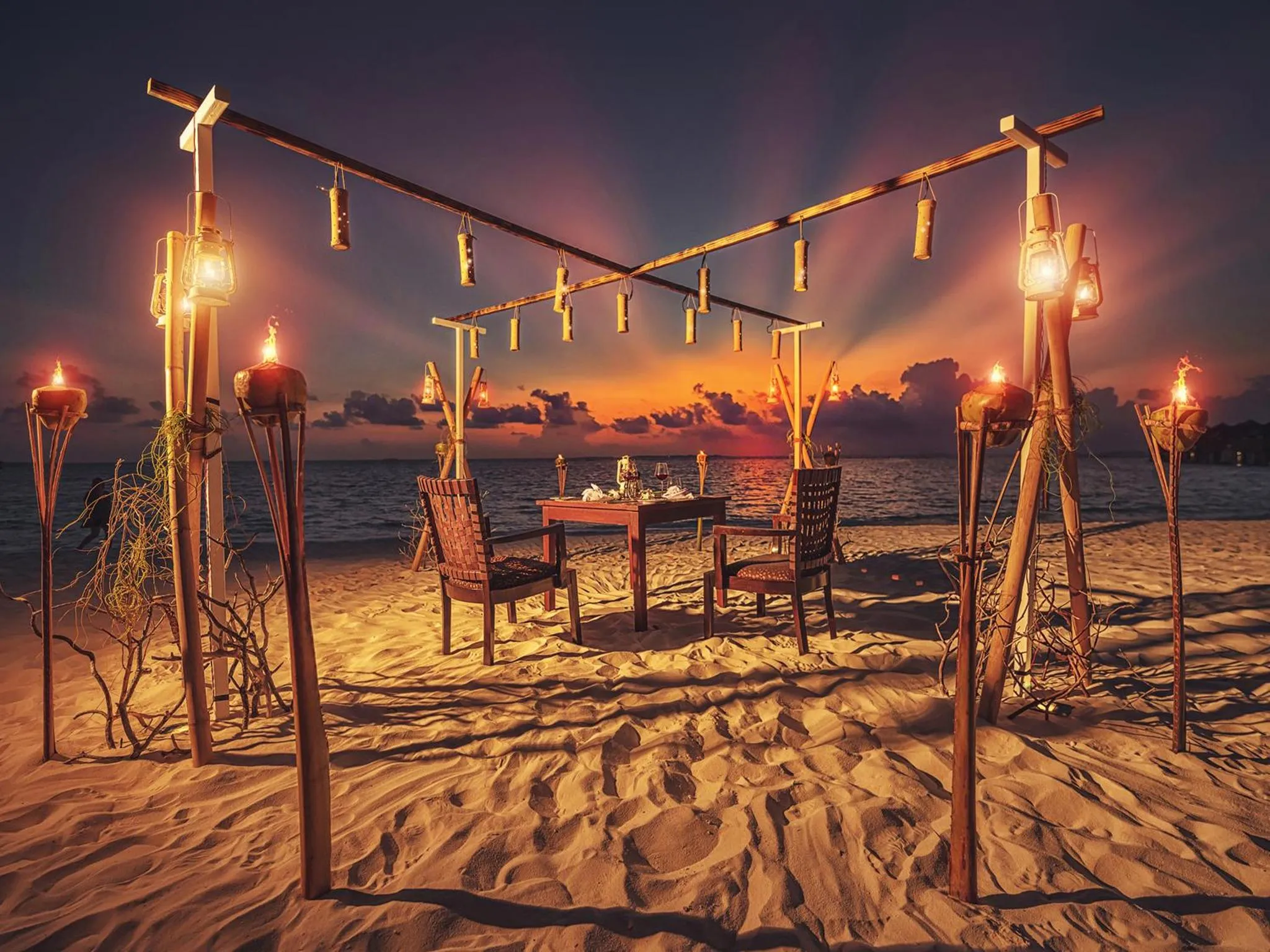 Table romantique dressée sur une plage au coucher du soleil, éclairée par des lanternes.