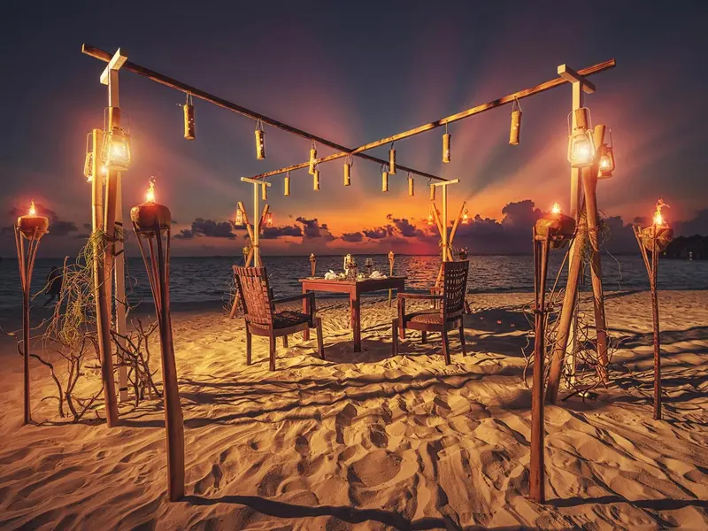 Table romantique dressée sur une plage au coucher du soleil, éclairée par des lanternes.