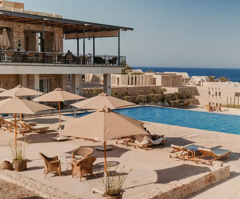 Transats et parasols bordent une piscine turquoise avec vue sur la mer.