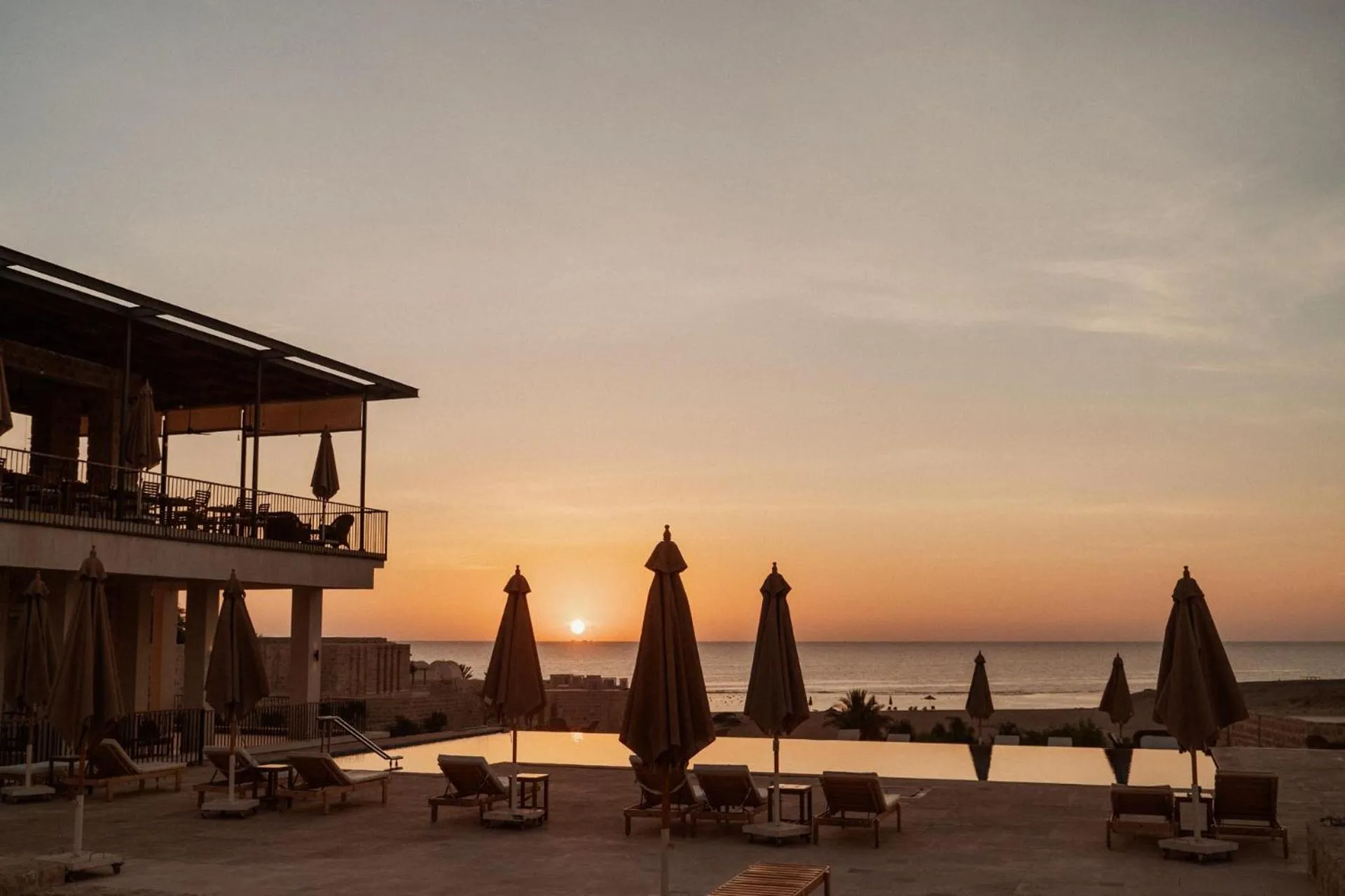Terrasse avec transats et parasols au bord d'une piscine face à la mer au coucher du soleil.