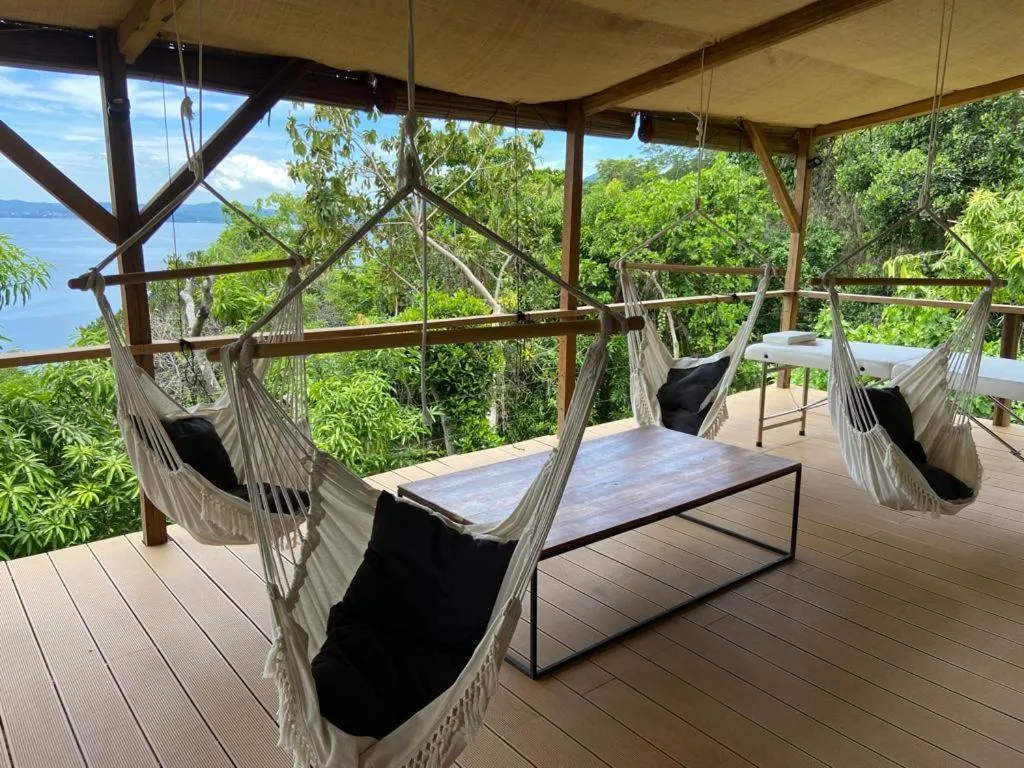 Terrasse en bois avec des hamacs et une table basse offrant une vue sur la mer et la végétation.