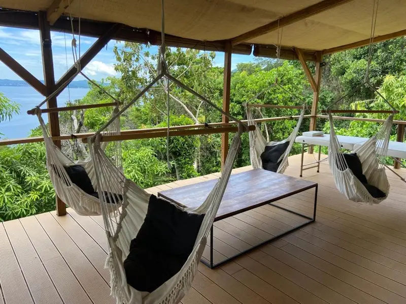 Terrasse en bois avec des hamacs et une table basse offrant une vue sur la mer et la végétation.