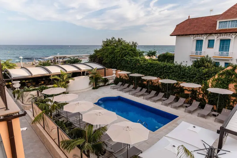 Vue aérienne d'une piscine entourée de transats et de parasols, avec un bâtiment blanc en arrière-plan et la mer au loin.