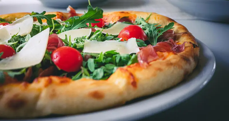 Une pizza garnie de tomates cerises, de roquette et de copeaux de parmesan est présentée sur une assiette.