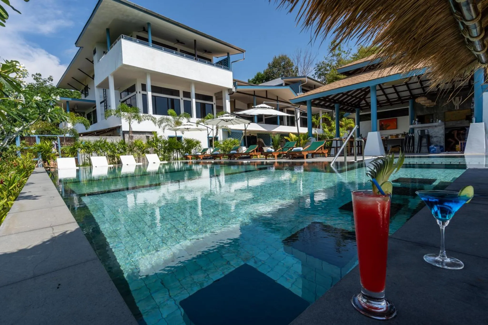 Une piscine turquoise bordée de transats, avec un bâtiment blanc en arrière-plan et des cocktails au premier plan.