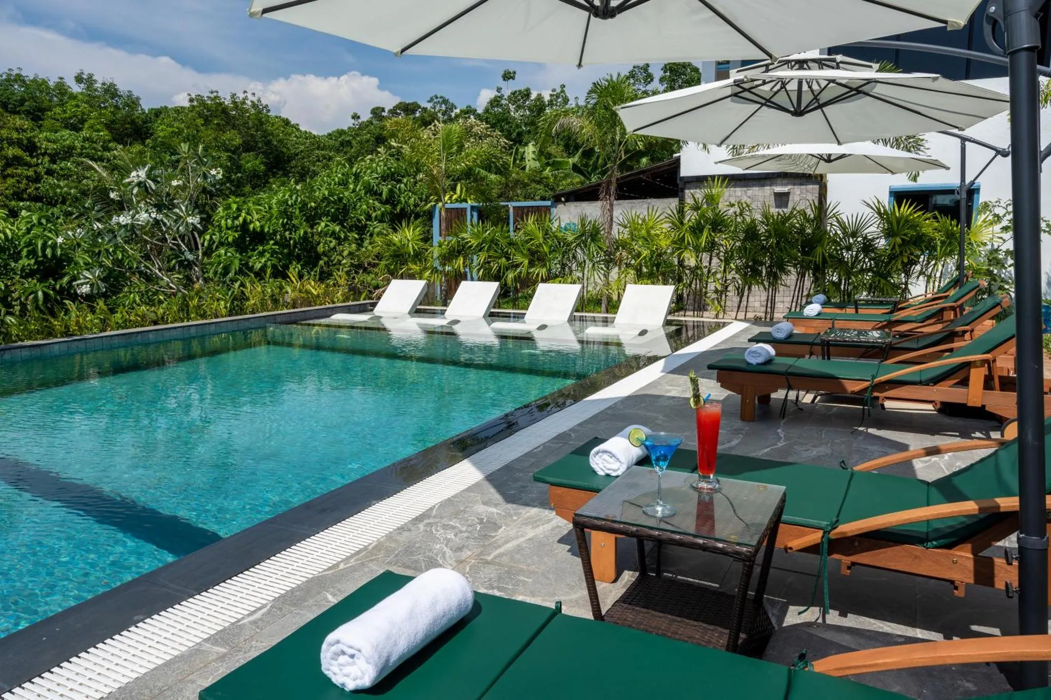 Transats verts et bleus bordent une piscine turquoise avec un cocktail rafraîchissant posé sur une table.