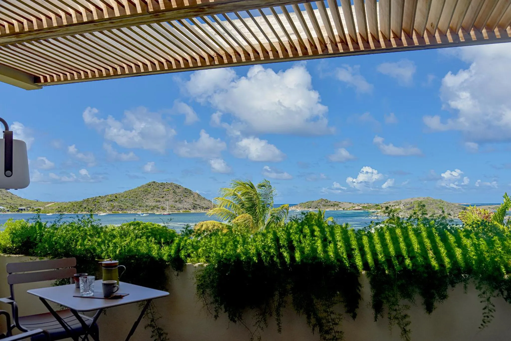 Une table avec des tasses sur une terrasse ombragée offre une vue sur la mer et la végétation.