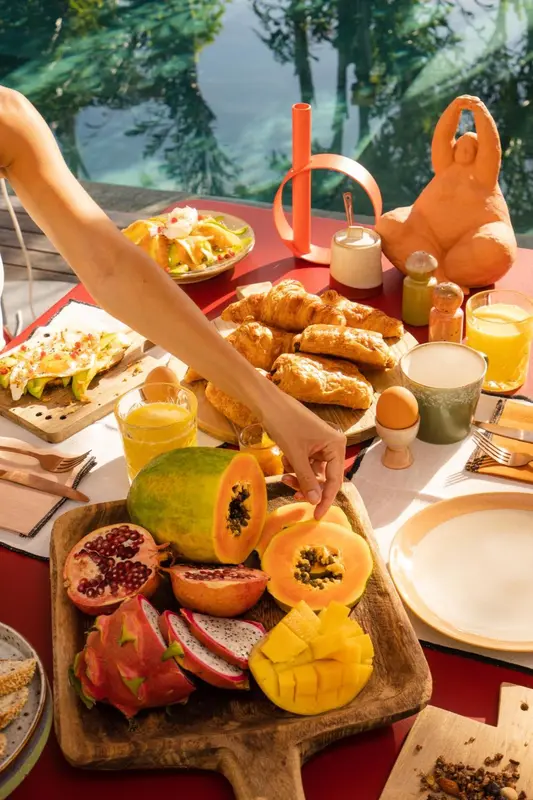 Une table colorée est dressée avec un copieux petit-déjeuner de fruits frais, viennoiseries et boissons.
