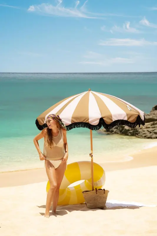 Une femme se tient sous un parasol rayé sur une plage de sable fin.