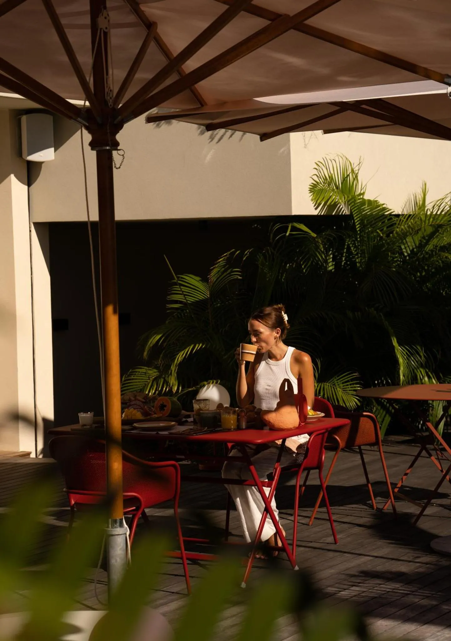 Une femme assise à une table rouge boit une boisson sur une terrasse ombragée.