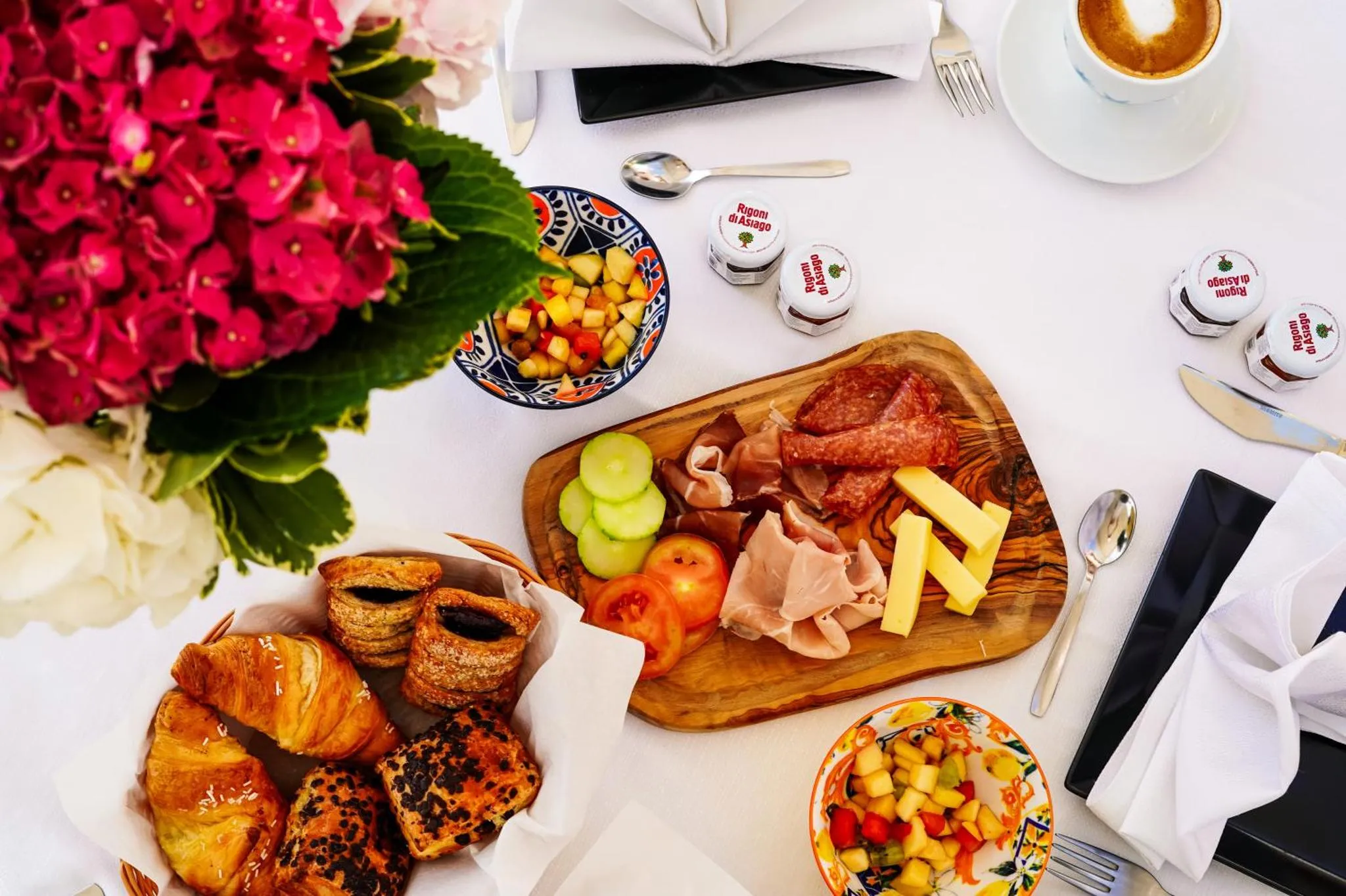 Une table est dressée avec un petit-déjeuner composé de viennoiseries, de fruits, de charcuterie et de boissons.