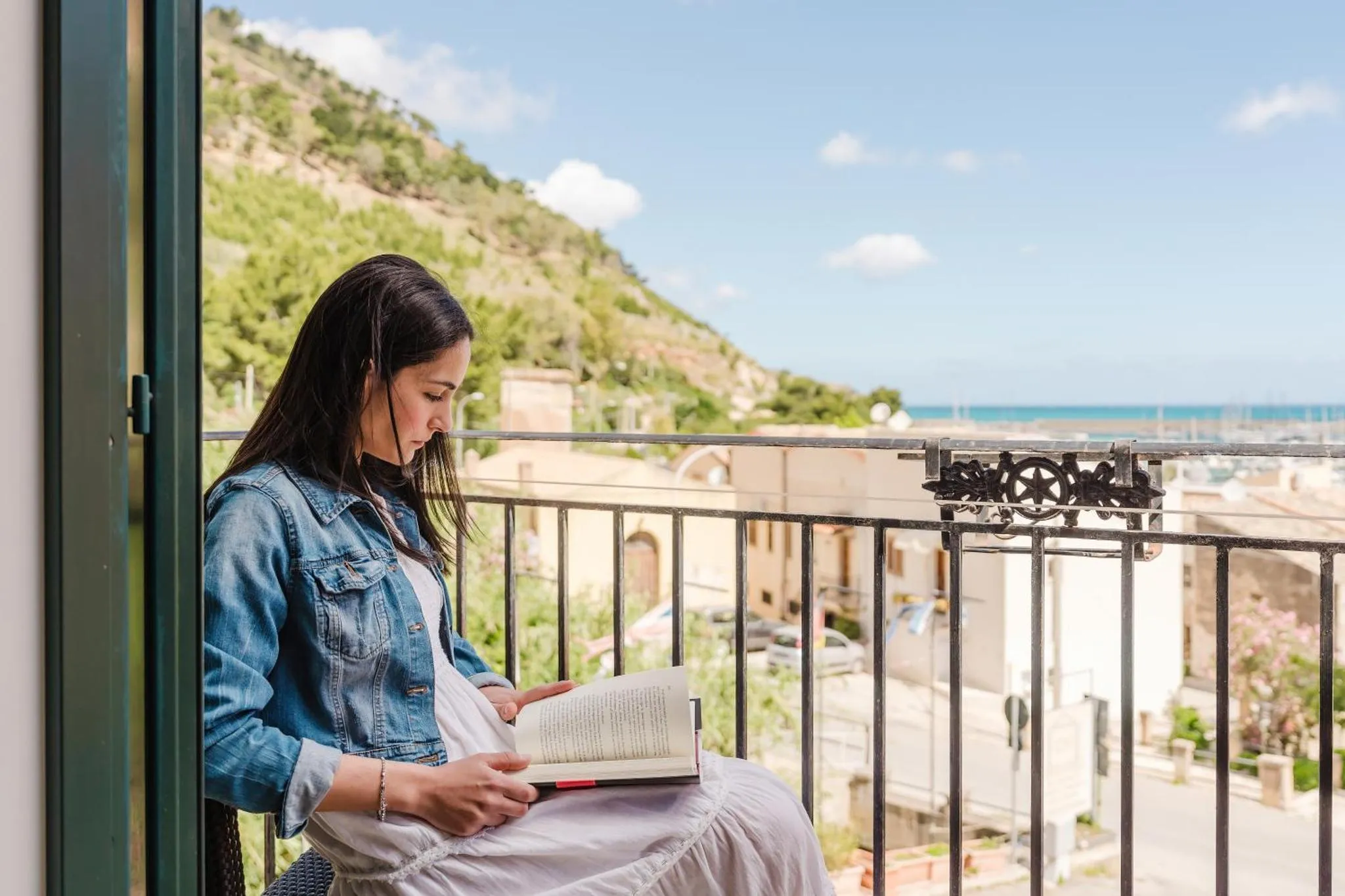 Une femme lit un livre assise sur un balcon avec vue sur la mer et les montagnes.