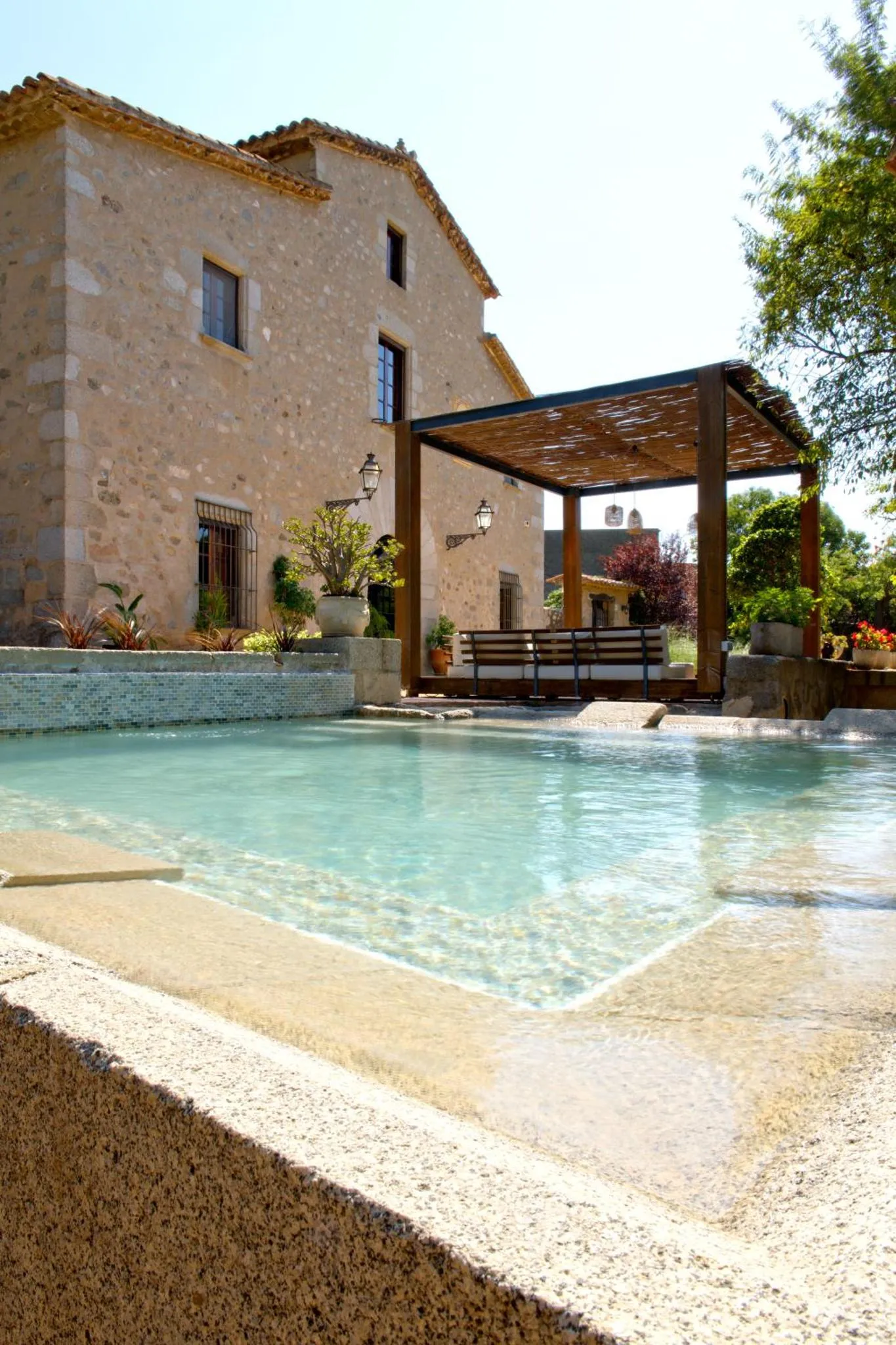Une piscine avec bordure de sable et une pergola ombragée.