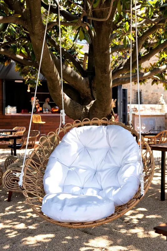 Un fauteuil suspendu en osier blanc se trouve sous un arbre, près d'un bar extérieur.