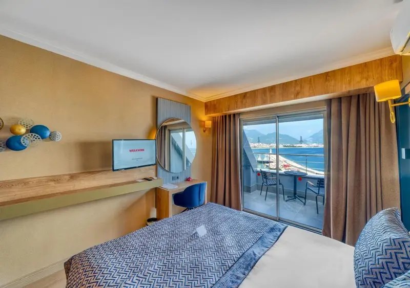 Une chambre lumineuse avec un lit recouvert d'une courtepointe bleue et une vue sur la mer depuis un balcon.