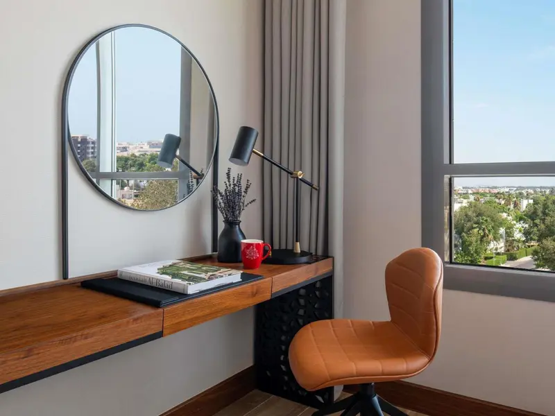 Un bureau en bois avec une lampe et un miroir rond près d'une fenêtre.