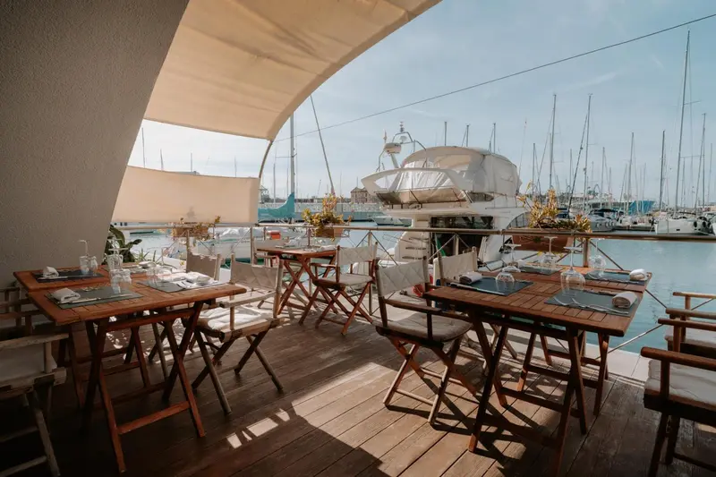 Tables et chaises en bois disposées sur une terrasse ensoleillée avec vue sur un port de plaisance.