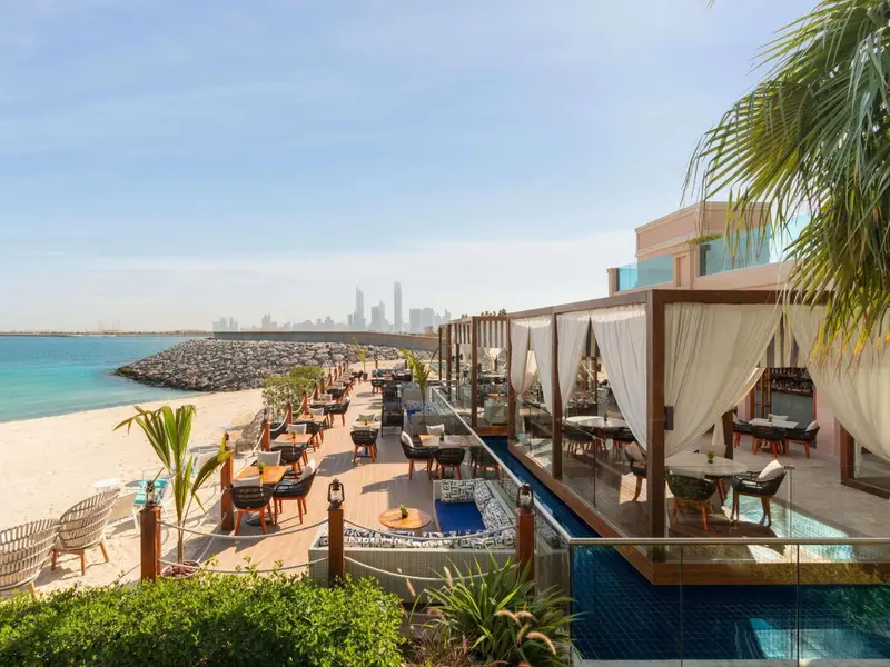 Terrasse ombragée avec tables et chaises donnant sur une plage et la mer.