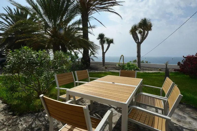 Une table et des chaises en bois clair sont disposées sur une terrasse ensoleillée, entourées de végétation luxuriante.