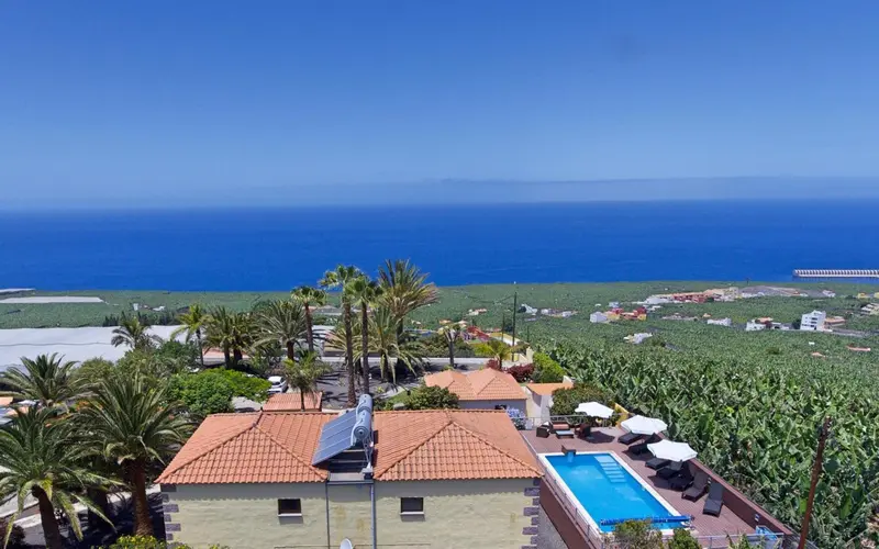 Vue aérienne sur des bâtiments aux toits rouges, une piscine et la mer au loin.
