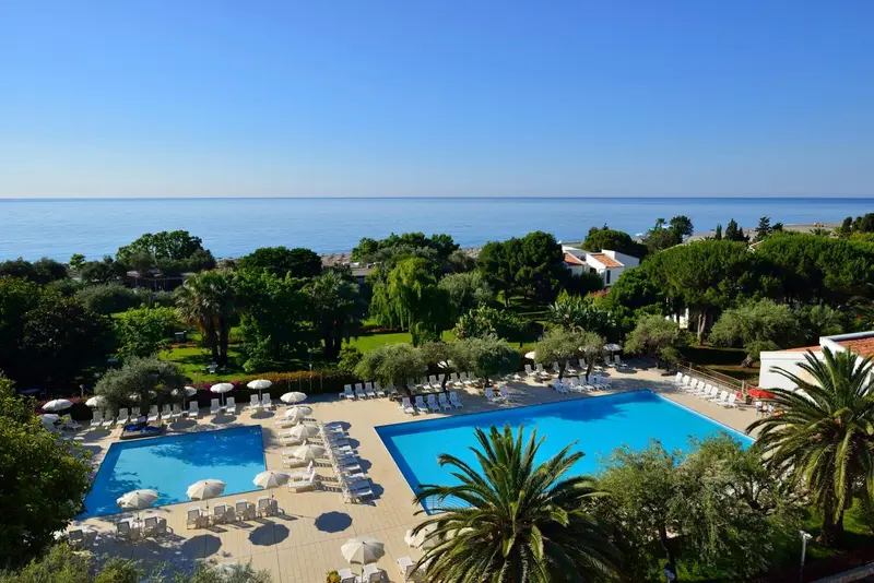 Vue aérienne d'une piscine entourée de transats et de végétation luxuriante, avec la mer en arrière-plan.