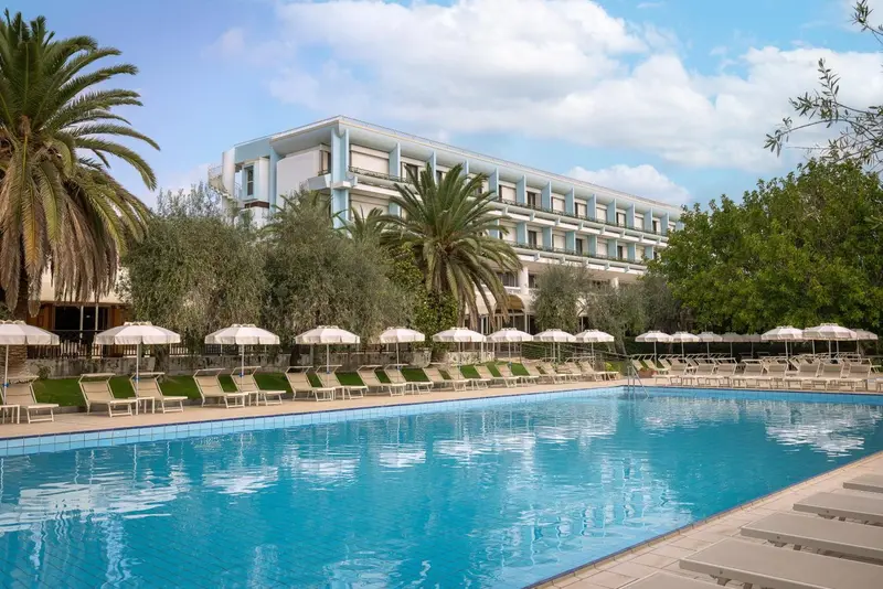 Une piscine turquoise bordée de transats et de parasols devant un bâtiment à plusieurs étages.