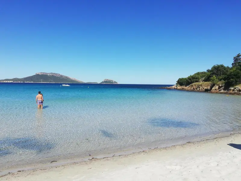 Une personne se tient dans une eau turquoise peu profonde sur une plage de sable clair.