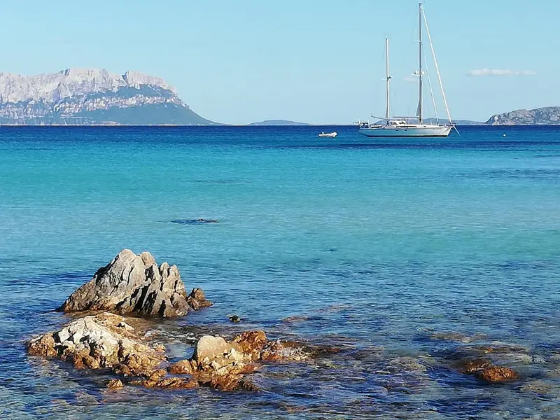 Vue sur une mer turquoise avec des rochers au premier plan et un voilier à l'horizon.