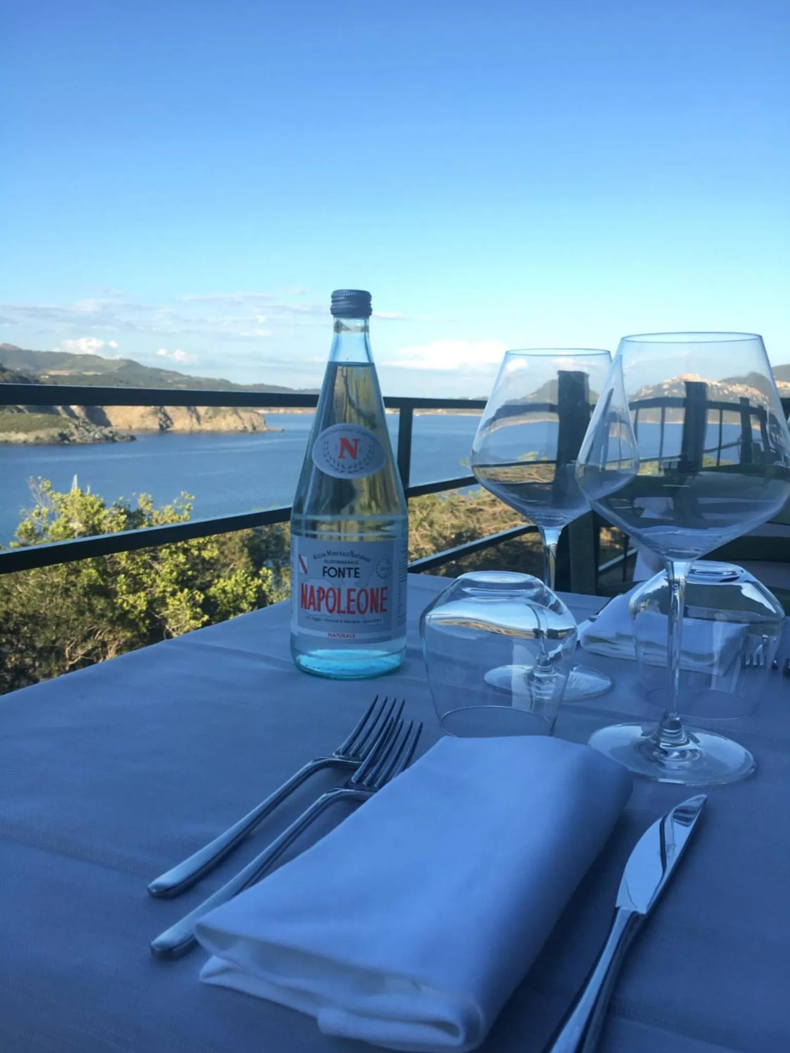 Une table dressée avec couverts et boissons offre une vue sur la mer.