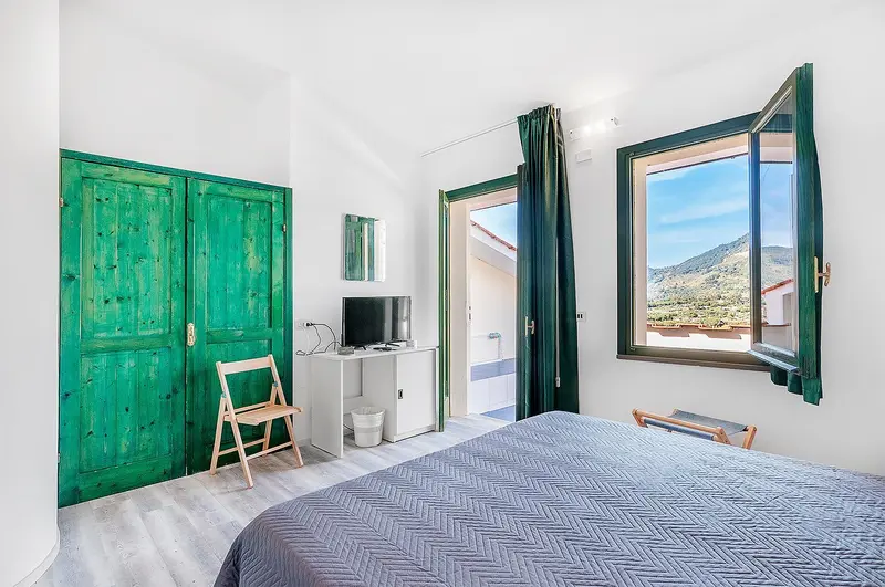 Une chambre lumineuse avec un lit fait et un bureau près d'une fenêtre ouverte sur un paysage montagneux.