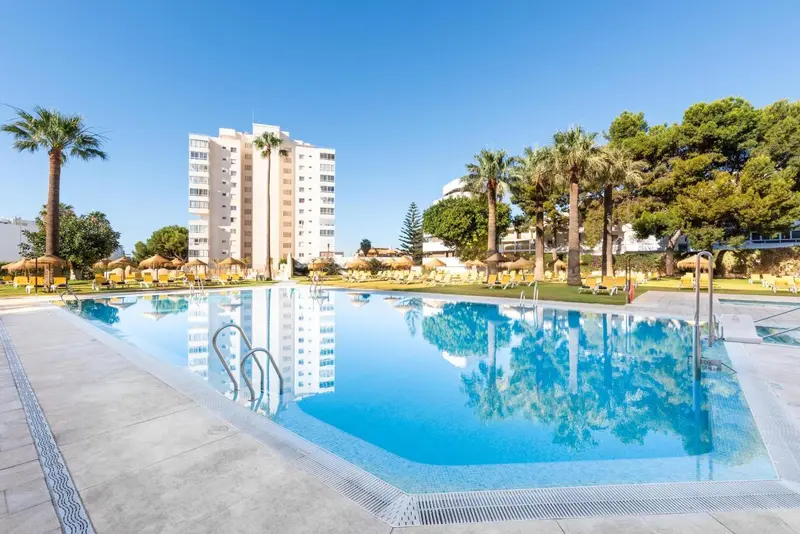 Une piscine rectangulaire reflète un bâtiment blanc et des palmiers sous un ciel bleu.