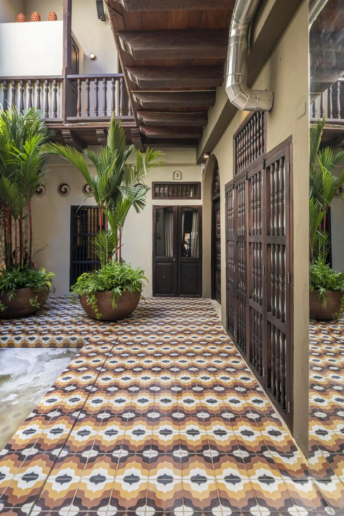 Une cour intérieure ornée de carreaux colorés et de plantes tropicales mène à une entrée sombre.