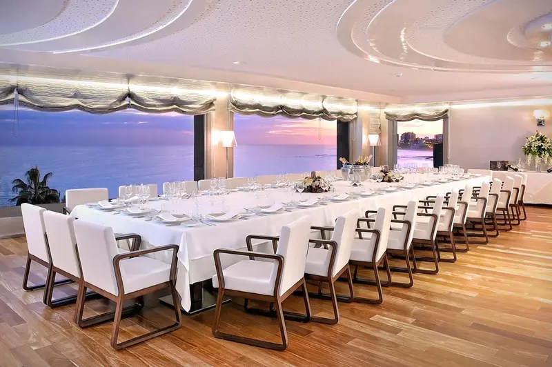 Une longue table est dressée pour un repas dans une salle lumineuse avec vue sur la mer.