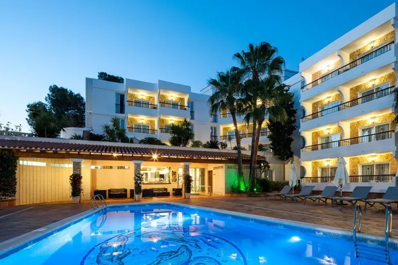 Bâtiment blanc illuminé avec piscine au premier plan et palmiers.