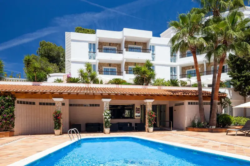 Bâtiment blanc à plusieurs étages avec une piscine au premier plan et des palmiers.