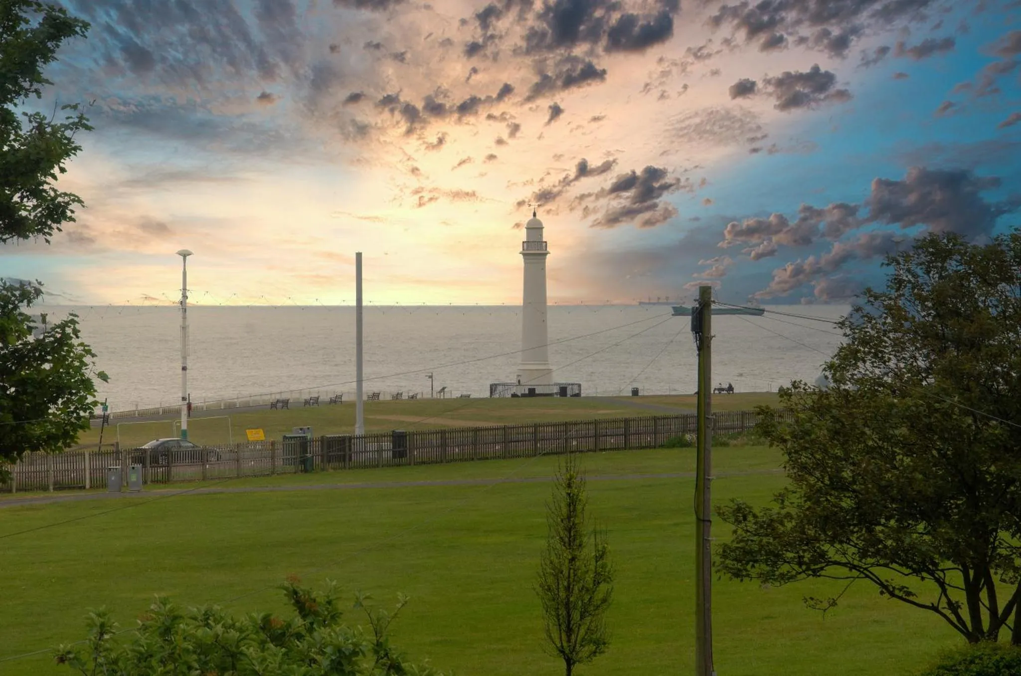 Un phare blanc se dresse au bord de la mer sous un ciel nuageux au coucher du soleil.