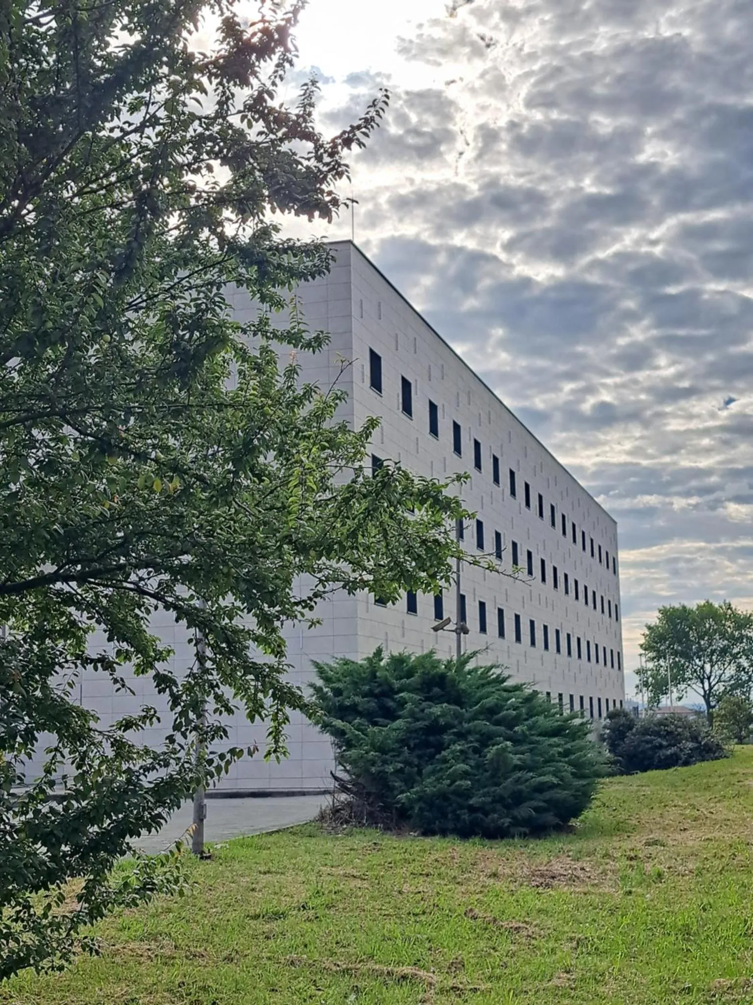 Bâtiment moderne blanc avec de nombreuses fenêtres, partiellement caché par la végétation.