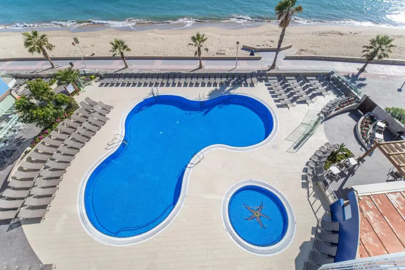 Vue aérienne d'une piscine d'hôtel bordée de transats, avec la plage en arrière-plan.