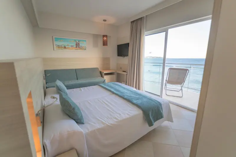Une chambre lumineuse avec un lit fait et un canapé turquoise, donnant sur une terrasse avec vue sur la mer.