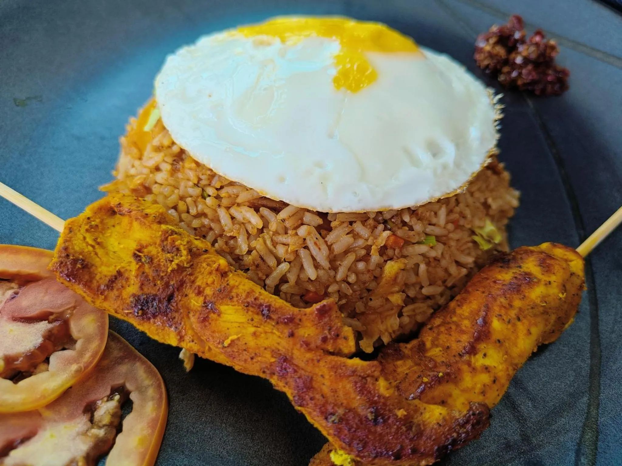 Assiette de riz frit surmontée d'un œuf au plat et accompagnée de brochettes de poulet et de légumes.
