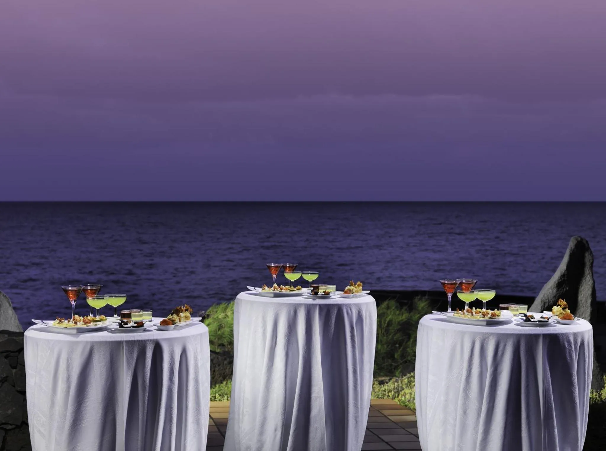 Tables dressées avec des plats et des boissons face à la mer au coucher du soleil.