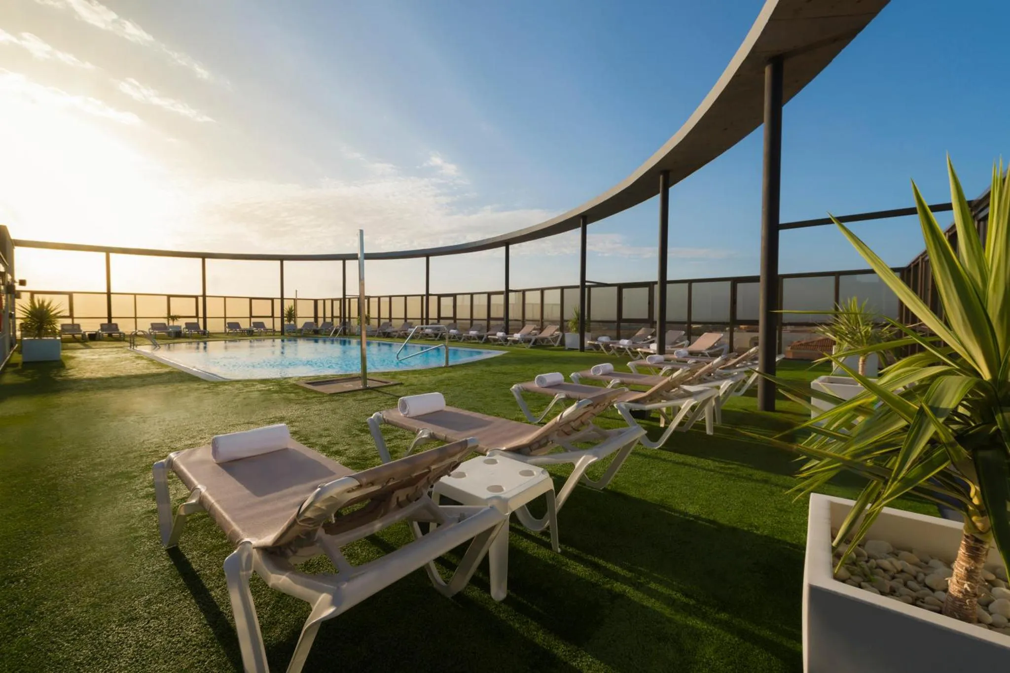 Transats blancs disposés autour d'une piscine rectangulaire sur une terrasse en gazon synthétique.