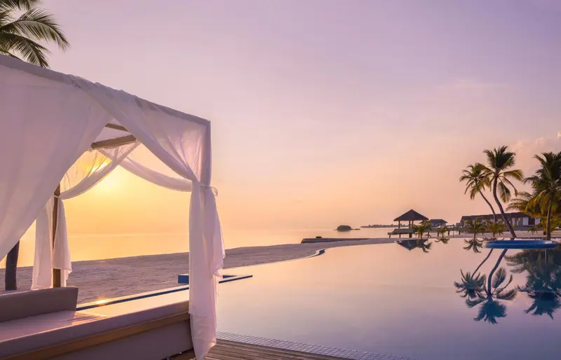 Un lit de plage ombragé surplombe une piscine à débordement face à l'océan au coucher du soleil.