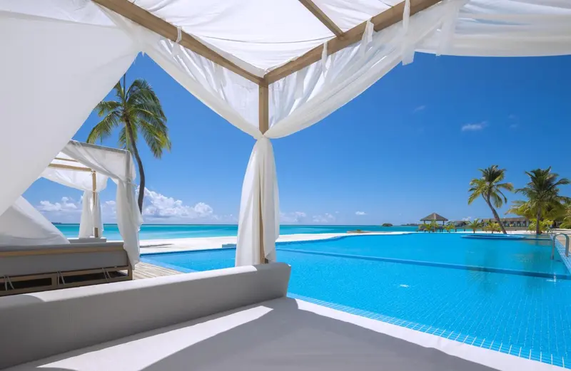 Transats blancs sous un auvent blanc surplombant une piscine turquoise et une plage.