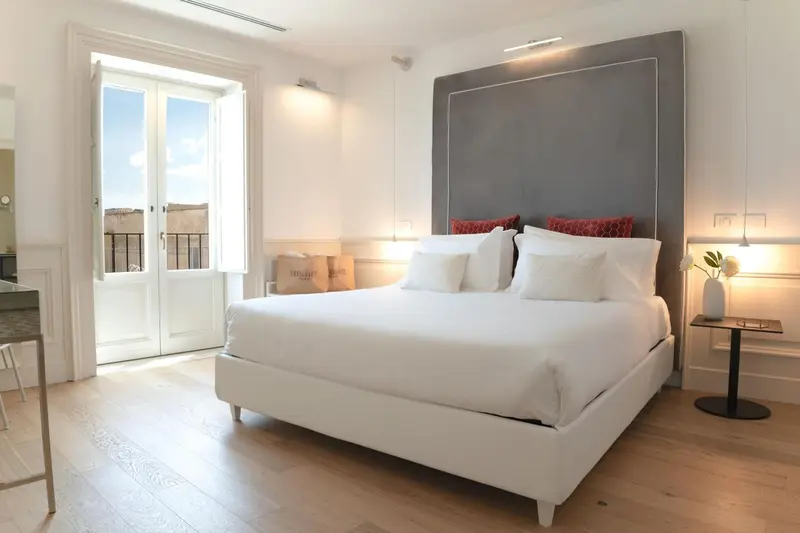 Une chambre lumineuse avec un grand lit blanc et des fenêtres ouvertes sur une vue extérieure.