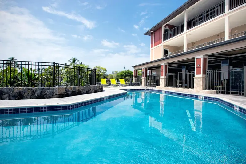 Piscine bleue bordée de chaises longues devant un bâtiment à balcons.
