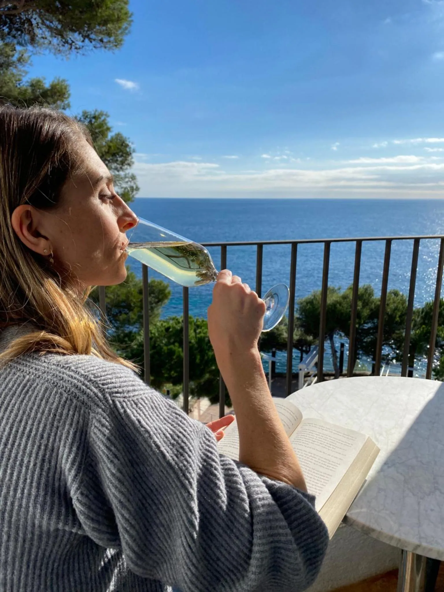 Une femme déguste une boisson sur une terrasse ensoleillée avec vue sur la mer.