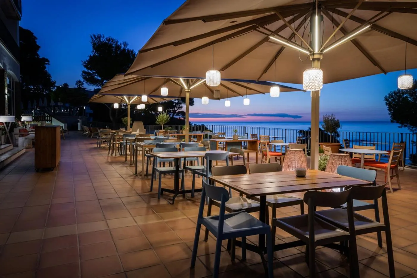 Terrasse ombragée avec tables et chaises donnant sur la mer au crépuscule.